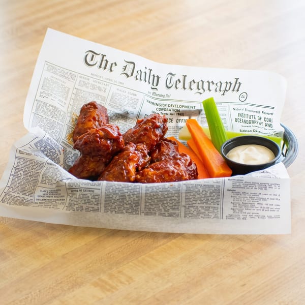 A basket of buffalo chicken wings, celery, carrots, and ranch dressing, served on newspaper.