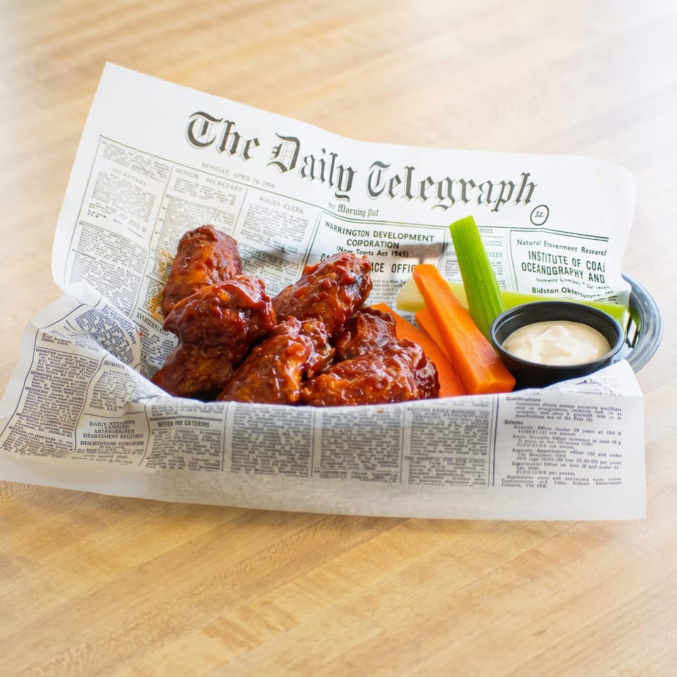 A basket of buffalo chicken wings, celery, carrots, and ranch dressing, served on newspaper.