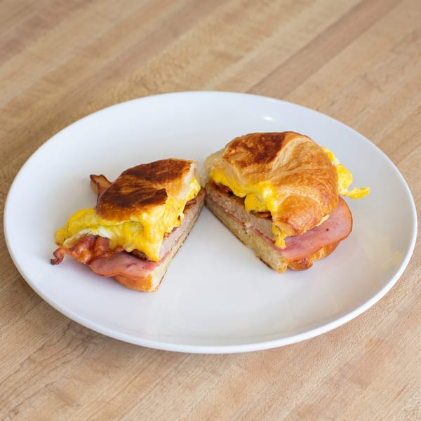 A croissant breakfast sandwich, cut in half, filled with scrambled eggs, bacon, and ham on a white plate.