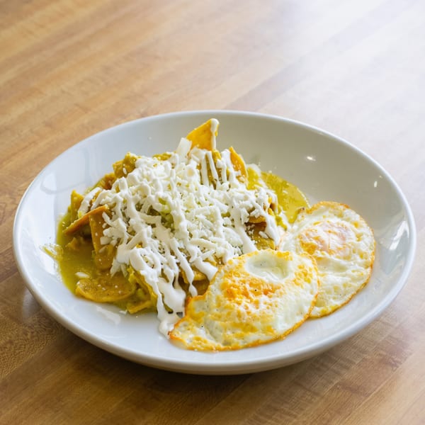 Chilaquiles with green sauce, shredded cheese, crema, and two fried eggs on a white plate.